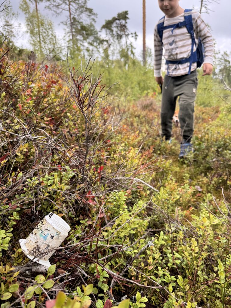 Barn med tursekk finner et forsøplet yoghurtbeger blant lyng og kvister i skogen.